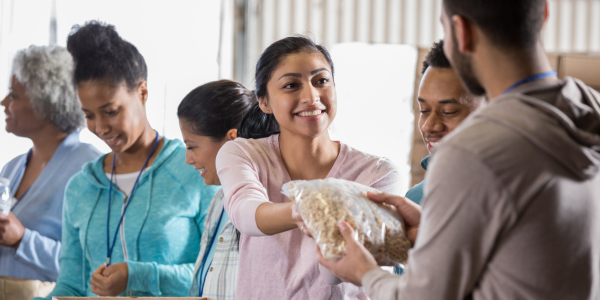A group of people in a room volunteering in a food pantry. The focus is on a woman in the middle who is wearing pink and smiling while handing a bag of food to a man in a beige hoodie.