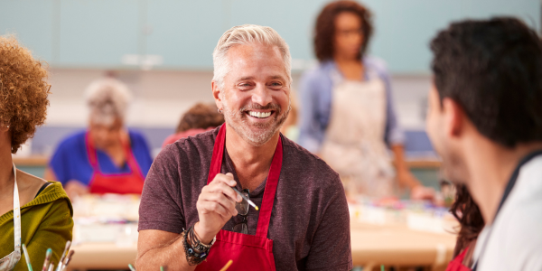 A middle-aged man wearing a grey top and red apron is smiling while holding a paintbrush He is in a painting group. There are other people in the background who are also part of the group.