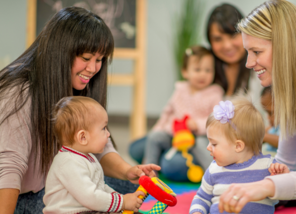 Two mothers with their children facing each other in a supported playgroup. The babies are playing with colourful toys.