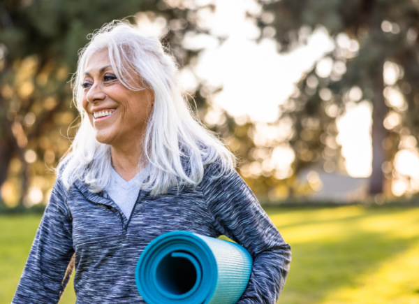 An older woman with grey hair is outdoors holdng a pilates mat and smiling. Behind her is a grass land.