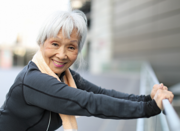 An older woman with grey hair smiling and stretching on a hand rail outdoors wearing a grey sports jacket.