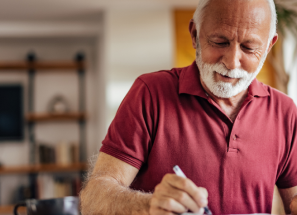 Older man in a red tshirt writing indoors on a book.