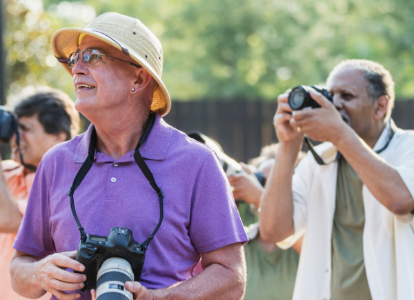 3 people taking photos outdoors using cameras. The man in the middle and in focal point is wearing purple and a sun hat while looking off to the distance. The man on the far left is focusing and wearing a salmon pink tshirt. The man on the right is also focusing on taking a photo and is wearing a white and green outfit.