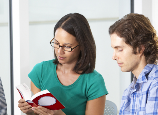 Two people indoors in a class. The woman in the centre is wearing turquoise and is reading a red book. The man on the far right is also looking down and focusing on a book that is off screen.