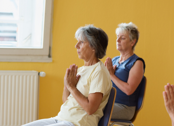 Women doing strength and balance exercises while seated on chairs. There is two older women on frame and they are in front of a yellow wall..