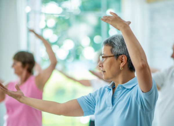 3 people in a Tai Chi class doing the same pose which involves stretching out the right up and putting the left arm in the air. The woman in the middle is wearing blue and has short hair. The woman behind her is wearing pink. The man behind her on the right of the image is wearing grey,