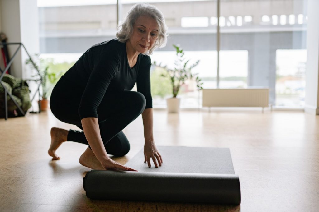 Smiling woman with grey hair bends down to adjust her yoga mat